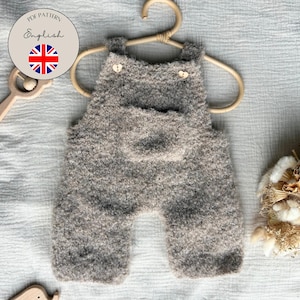 Könnte beinhalten: Ein flauschiger, grauer Strick-Strampler für Babys mit einer Vordertasche und zwei kleinen herzförmigen Knöpfen an den Schulterträgern. Das Kleidungsstück hängt an einem hellen Holzkleiderbügel. Ein rundes Abzeichen trägt den Text PDF PATTERN English.