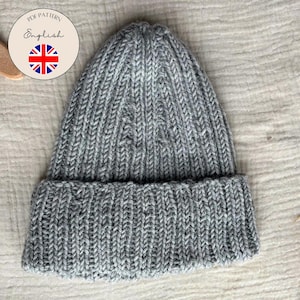 Könnte beinhalten: Graue Strickmütze mit umgeschlagenem Saum. Die Mütze ist aus strukturiertem Garn gefertigt und hat ein Rippenmuster. Eine runde Grafik mit dem Text "PDF Pattern English" und einer Union Jack-Flagge ist sichtbar.