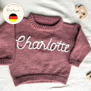 Op de afbeelding: Een stoffig roze gebreide trui met de naam "Charlotte" in het wit geborduurd. De trui heeft een geribbelde kraag, manchetten en zoom. De afbeelding bevat ook houten speelgoed en tekst in het Duits.