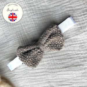 May include: A knitted brown bow tie with white fabric ends, likely for a baby or child. The bow tie is centered on a textured, light gray surface. A circular graphic with the text "PDF Pattern English" and a British flag is in the upper left corner.