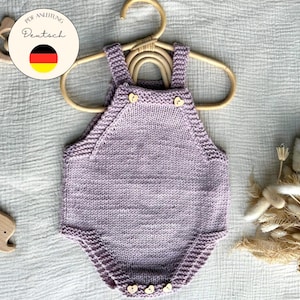 Könnte beinhalten: Ein fliederfarbener gestrickter Baby-Strampler mit Holzknöpfen und Trägern. Der Strampler wird auf einem Holzbügel präsentiert. Ein Holz-Elefanten-Spielzeug und eine Holzform sind ebenfalls sichtbar.