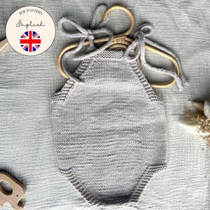 Könnte beinhalten: Hellgrauer gestrickter Baby-Strampler mit Neckholder, an einem Holzkleiderbügel hängend. Der Strampler ist aus weichem Garn gefertigt und hat ein strukturiertes Aussehen. Ein Holz-Elefanten-Spielzeug und ein rundes Etikett mit dem Union Jack sind ebenfalls sichtbar.