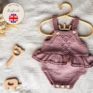 Può includere: Un body per neonati lavorato a maglia in rosa antico, con una gonna a balze e bottoni a forma di cuore. Il body è appeso a un appendiabiti di legno. Sono visibili anche giocattoli di legno, tra cui un sonaglio e un elefante.