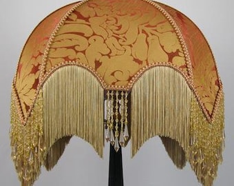 May include: A gold and orange lampshade with a fringe trim and beaded accents. The lampshade is made of fabric with a damask pattern.