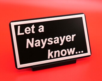 Puede incluir: Un letrero negro con un borde blanco y texto blanco que dice "Let a Naysayer know..." El letrero está sobre una superficie roja.