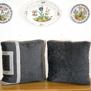 May include: Two dark gray square throw pillows with a textured appearance. One pillow features a light gray geometric design. Above, three decorative plates with floral patterns hang on a white wall.