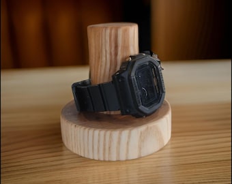 Puede incluir: Un reloj digital negro con una banda de goma negra se encuentra sobre un soporte de reloj de madera. La esfera del reloj tiene el texto "G-SHOCK" impreso en él.
