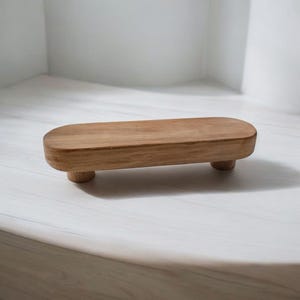 May include: A small, oval-shaped wooden tray with rounded edges and three legs. The tray is made of light brown wood and has a smooth, polished finish.