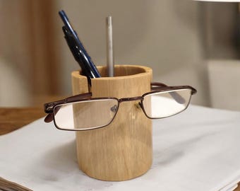 May include: A wooden pencil holder with a slot for glasses. The holder is made of light brown wood and has a natural finish. Two pens and a pencil are in the holder. A pair of glasses is resting on the slot.