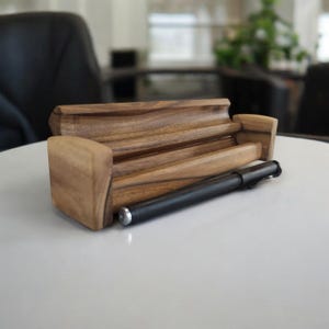 May include: A wooden pen holder with a natural wood finish. The holder has two compartments and is designed to hold pens or pencils. A black pen is resting on top of the holder.