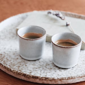 Könnte beinhalten: Zwei kleine, weiße Keramikbecher mit Espresso stehen auf einem gesprenkelten, cremefarbenen Keramiktablett. Die Tassen haben einen braunen Rand und der Espresso hat eine satte, dunkle Farbe. Ein kleines Buch und eine Trockenblume befinden sich ebenfalls auf dem Tablett.