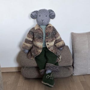 May include: A gray knitted sheep doll wearing a brown and green striped sweater and green pants. The doll is sitting on a brown cushion.