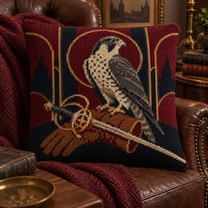 May include: A square knit throw pillow featuring a detailed graphic of a falcon perched on a brown leather falconry glove. The design includes a sword, a dark red circular background, and architectural silhouettes in deep navy blue and dark red.