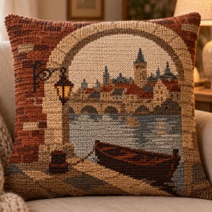 Può includere: Un cuscino quadrato con una dettagliata scena a mezzo punto raffigurante un paesaggio urbano europeo al crepuscolo. Il design include un arco in pietra, un lampione acceso, un fiume e una piccola barca in legno ormeggiata a un molo, nei toni del marrone, beige e blu.