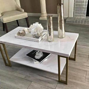 May include: A white marble-topped coffee table with gold-colored metal legs. The table has a lower shelf and is decorated with vases, a tray with decor, and a book. The table is 48 inches long and 24 inches wide.