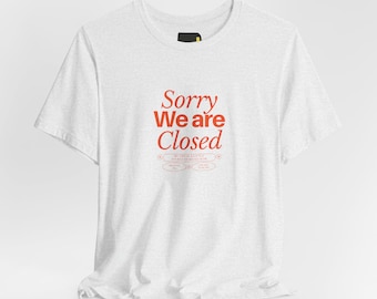 May include: A light grey short-sleeve t-shirt featuring the red text Sorry We are Closed printed on the centre chest. Below the main text, smaller red text and oval shapes are visible, including the phrase My brain is a little flooded right now.