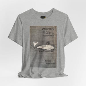 Op de afbeelding: Een grijs gemêleerd t-shirt met korte mouwen en een rechthoekige grafische print van een witte walvis in het water. De tekst luidt Moby Dick or The White Whale by Herman Melville. Het ontwerp heeft een textuur die lijkt op canvas.