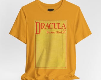 May include: A mustard yellow short-sleeve t-shirt featuring a rectangular graphic on the chest. The graphic contains the red text "Dracula By Bram Stoker" at the top, framed by a thin red border against a textured light yellow background.