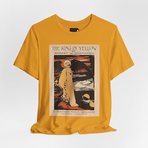 Op de afbeelding: Een mosterdgeel t-shirt met korte mouwen en een rechthoekige grafische print van de boekomslag The King in Yellow door Robert W. Chambers. De print toont een figuur met vleugels in een donker landschap en bevat bijbehorende tekst.