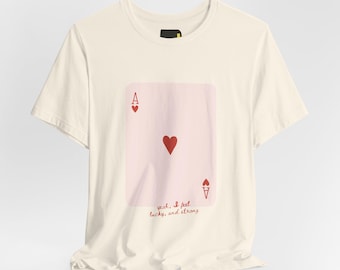 May include: A cream-coloured short-sleeve t-shirt featuring a large graphic of a pink ace of hearts playing card on the chest. The graphic includes the text 'yeah, I feel lucky, and strong' in red cursive script below the central heart symbol.