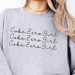 以下が含まれることがあります： 手書きのフォントで「Coke Zero Girl」という文字が3回繰り返されたグレーのトレーナーです。
