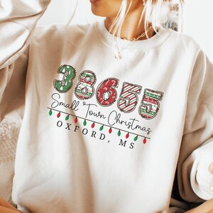 May include: A cream colored sweatshirt with a festive design featuring the numbers 365 with red and green accents. The text "Small Town Christmas" is written below the numbers, and "Oxford, MS" is written below the text.