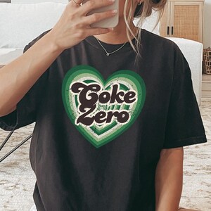 以下が含まれることがあります： レトロなフォントで「Coke Zero」と書かれた、緑と白のハートのグラフィックが描かれた黒いTシャツです。