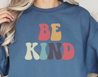 Könnte beinhalten: Ein blaues Sweatshirt mit den Worten "BE KIND" in farbenfrohen Buchstaben im Retro-Stil. Der Text ist in Rot, Gelb, Blau, Grün und Lila gehalten. Das Sweatshirt hat eine einfarbige blaue Farbe.