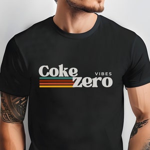 Puede incluir: Camiseta negra con un gráfico de estilo retro que dice "Coke Zero Vibes" en texto blanco con rayas de colores.