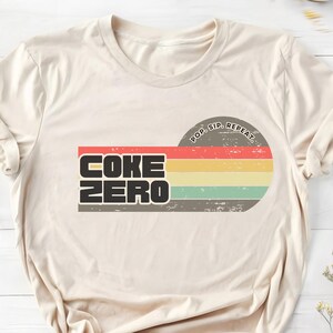 以下が含まれることがあります： クリーム色のTシャツ。レトロなデザインで、黒い太字で「COKE ZERO」の文字が描かれています。赤、黄、緑、青の横縞模様があります。「POP. SIP. REPEAT.」というフレーズがデザインの上にあります。