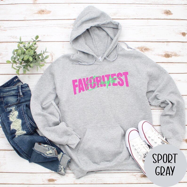 May include: A light grey hooded sweatshirt with the word "FAVORITEST" in pink and green. The sweatshirt has a front pocket and a drawstring hood. White trainers and blue jeans are next to the sweatshirt.