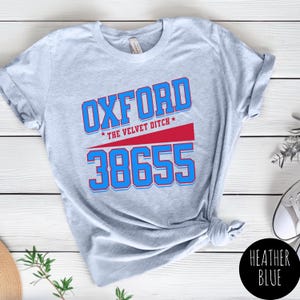 Puede incluir: Camiseta azul jaspeado con la palabra "OXFORD" en letras azules y rojas, el texto "THE VELVET DITCH" y el número "38655". La camiseta está anudada en la parte inferior y tiene las mangas dobladas.