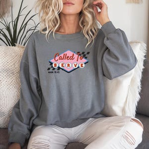 Könnte beinhalten: Graues Sweatshirt mit dem Schriftzug "Called To Serve" in einem Retro-Design. Das Design umfasst eine rosa Raute, ein schwarz-weißes Schachbrettmuster und den Text "Mark 10:45".