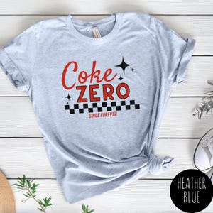 以下が含まれることがあります： 赤色の文字で「Coke Zero」と書かれた、黒と白の市松模様と「SINCE FOREVER」の文字が入った、ヘザーブルーのTシャツ。右下には「HEATHER BLUE」と書かれた黒い円があります。