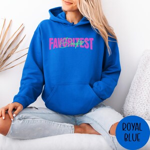 May include: Royal blue hoodie with the text "FAVORITEST Daughter" in pink and green. The hoodie features a front pocket and a drawstring hood. The image shows a person wearing the hoodie.