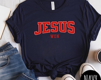 May include: Navy blue t-shirt with the words "JESUS WON" in red, collegiate-style lettering. The shirt is styled with blue jeans and white sneakers. The word "NAVY" is in a black circle.