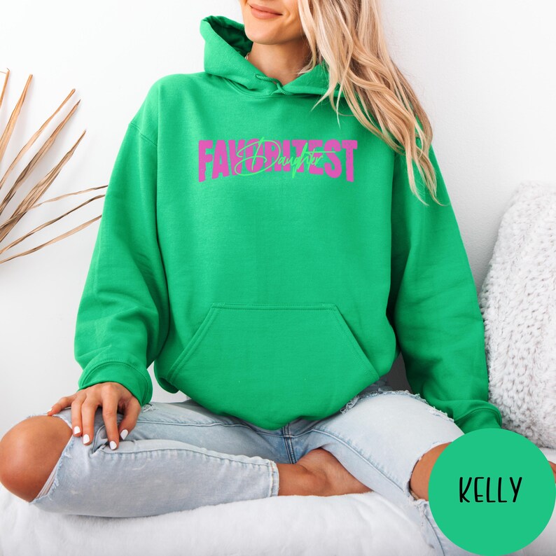 May include: A green hoodie with the text "FAV DAUGHTER" in pink and green. The sweatshirt has a front pocket and a hood. A green circle with the name "KELLY" is in the lower right corner.