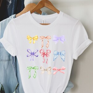 Könnte beinhalten: Weißes T-Shirt mit einem bunten Design aus neun verschiedenen Schleifen. Die Schleifen sind in verschiedenen Farben, darunter Gelb, Orange, Rosa, Lila, Blau und Grün.