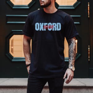 Op de afbeelding: Zwart T-shirt met ronde hals met het woord "OXFORD" in lichtblauwe blokletters over de borst. Onder het woord "OXFORD" staat het woord "22455" in rood. Het T-shirt wordt door een persoon gedragen.