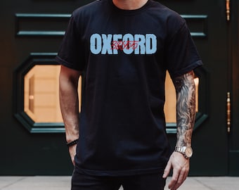 May include: Black crew neck t-shirt with the word "OXFORD" in light blue block letters across the chest. Below the word "OXFORD" is the word "22455" in red. The shirt is worn by a person.