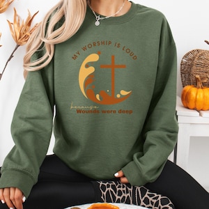 Könnte beinhalten: Ein olivgrünes Sweatshirt mit dem Text "MY WORSHIP IS LOUD" und "because Wounds were deep". Das Design umfasst ein Kreuz, Wellen und eine halbmondförmige Form in Orange- und Gelbtönen. Das Sweatshirt ist ein lässiger Stil.