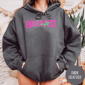 May include: Dark grey hoodie with a front pocket and a drawstring hood. The front displays the text "FAVORITEST DAUGHTER" in pink and green. A dark grey circle with the words "DARK HEATHER" is in the bottom right corner. A casual, comfortable garment.