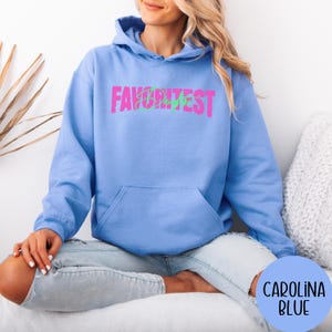May include: A Carolina blue hoodie with a front pocket and a drawstring hood. The hoodie displays the text "FAVORITEST" in pink and green. The words "Carolina Blue" are in a blue circle in the lower right corner.