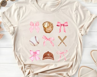 May include: A beige t-shirt with a baseball theme featuring pink bows, a baseball, a baseball glove, bats, and a baseball cap.