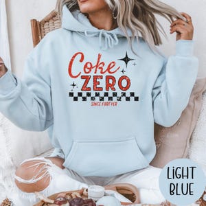 以下が含まれることがあります： フロントポケットとフードが付いた水色のパーカー。「Coke ZERO」の文字が赤色で、白と黒のチェック柄と「SINCE FOREVER」の文字がプリントされています。「LIGHT BLUE」の文字が水色の円の中にあります。