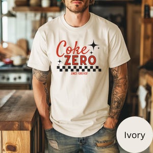 以下が含まれることがあります： アイボリー色のTシャツ。赤色の「Coke ZERO」の文字、白と黒の市松模様、そして「SINCE FOREVER」の文字がプリントされています。半袖でクルーネックです。