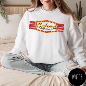 Puede incluir: Una sudadera blanca de manga larga con un gráfico en el pecho. El gráfico muestra una banda horizontal roja con efecto desgastado y una forma de letrero amarillo estilo marquesina que contiene el texto Oxford y The Velvet Ditch en negro.