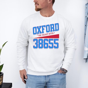 May include: A white long-sleeve sweatshirt with the word "OXFORD" in blue letters with a red stripe and the number "38655" in blue. The text "THE VELVET DITCH" is above the red stripe.