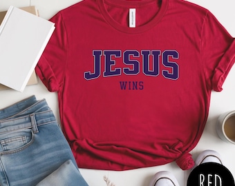May include: A red t-shirt with the text "JESUS WINS" in navy blue block letters. The shirt is paired with blue jeans and white sneakers. A black circle with the word "RED" is in the bottom right corner.