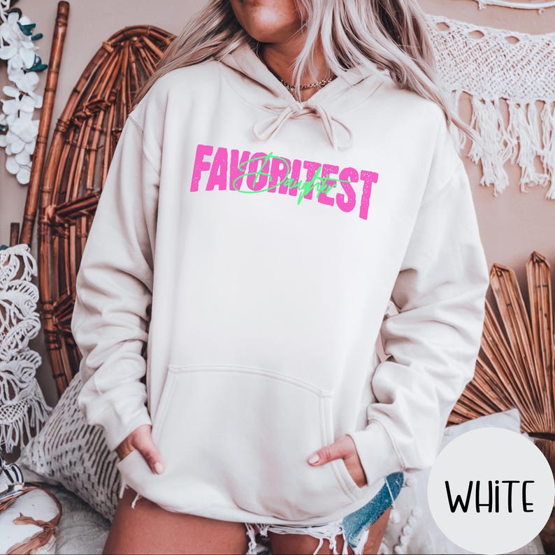 May include: A cream-coloured hoodie featuring the word "FAVORITEST" in pink block letters, with a green lightning bolt design. The hoodie has a front pocket and a drawstring hood. The word "WHITE" is in a white circle in the bottom right corner.